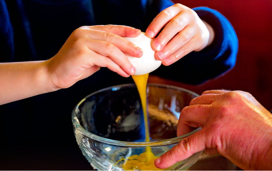 Mão de criança quebrando ovo na tigela, com mão de adulto segurando. Imagem ilustrativa texto receitas para o Dia dos Pais.