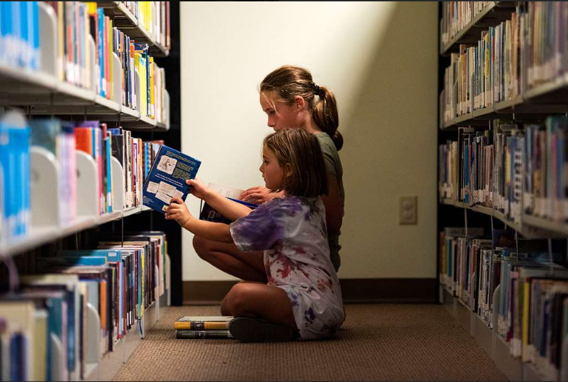 Duas meninas sentadas no chão, escolhendo livros na biblioteca. Imagem ilustrativa texto biblioteca atrativa.